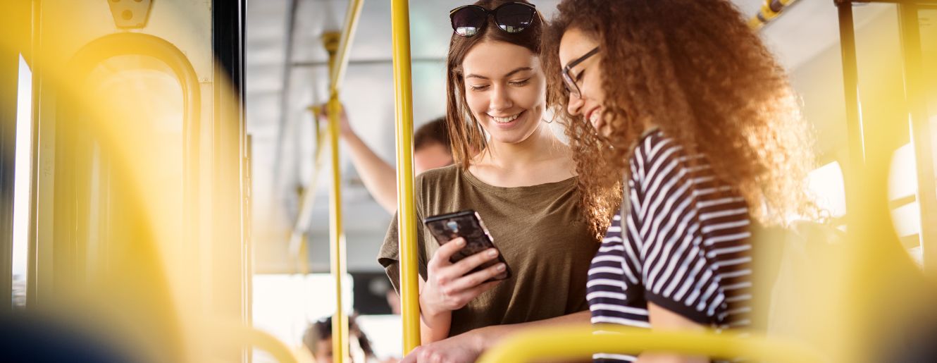 two female presenting people looking at a phone while standing on a bus. 